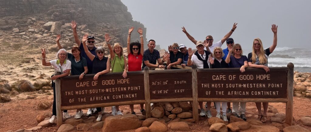 Reisegruppe mit hochgehobenen Armen hinter dem Schild am Kap der Guten Hoffnung